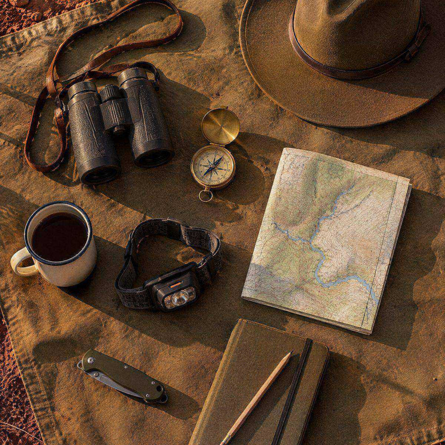 Field kit flat lay on canvas: binoculars, felt hat, compass, map, headtorch, enamel mug and notebook