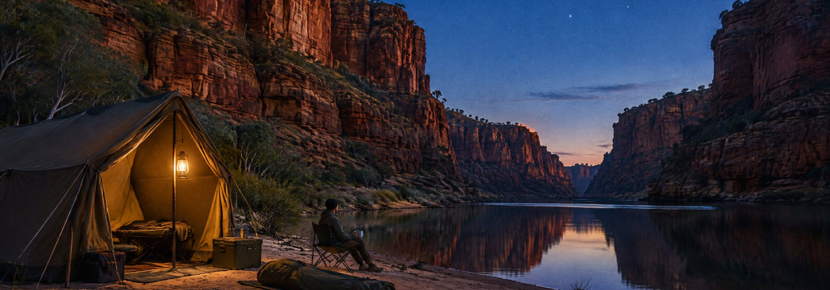 Canvas tent glowing at dusk beside a still river below red Kimberley gorge walls