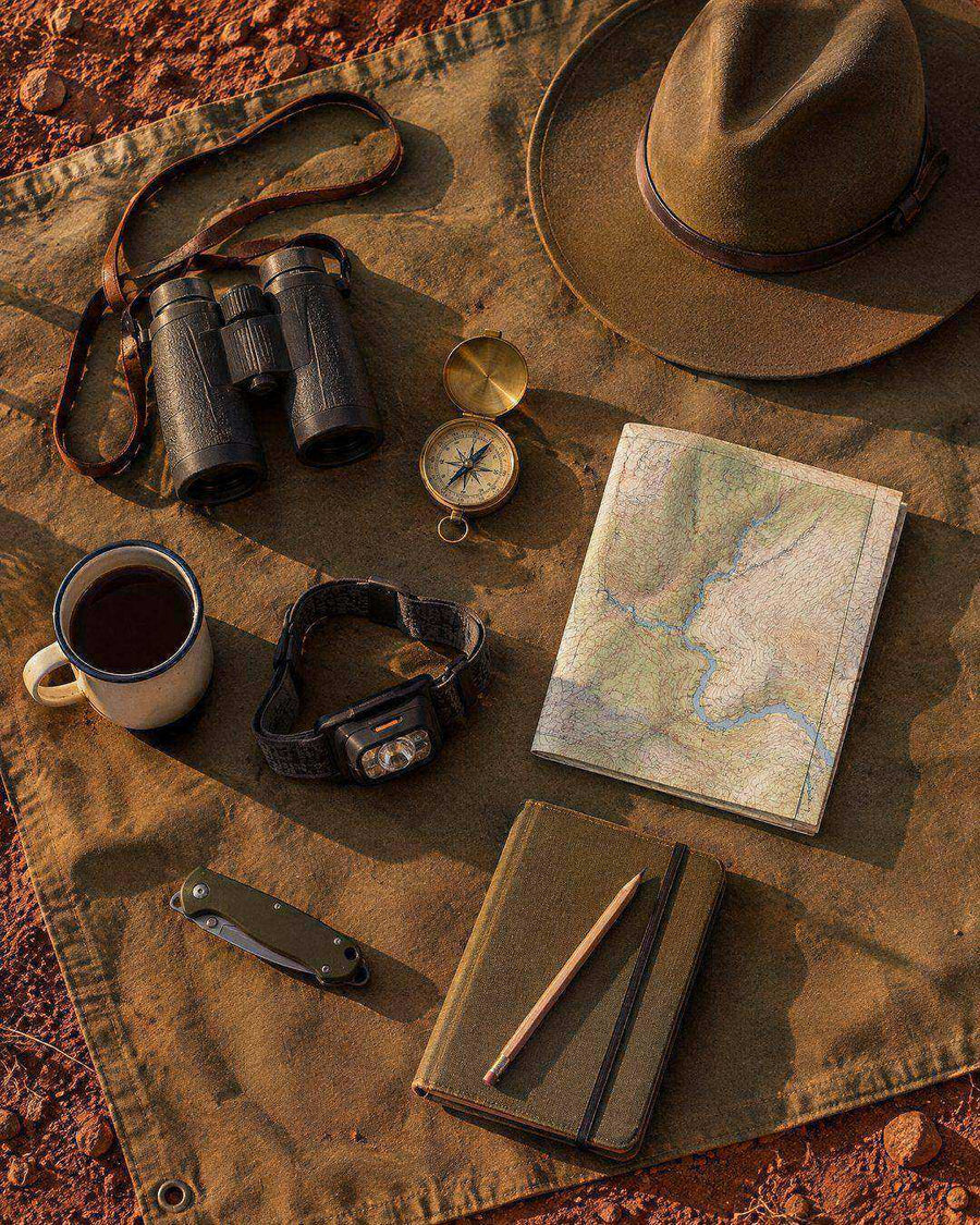 Field kit flat lay on canvas: binoculars, felt hat, compass, map, headtorch, enamel mug and notebook